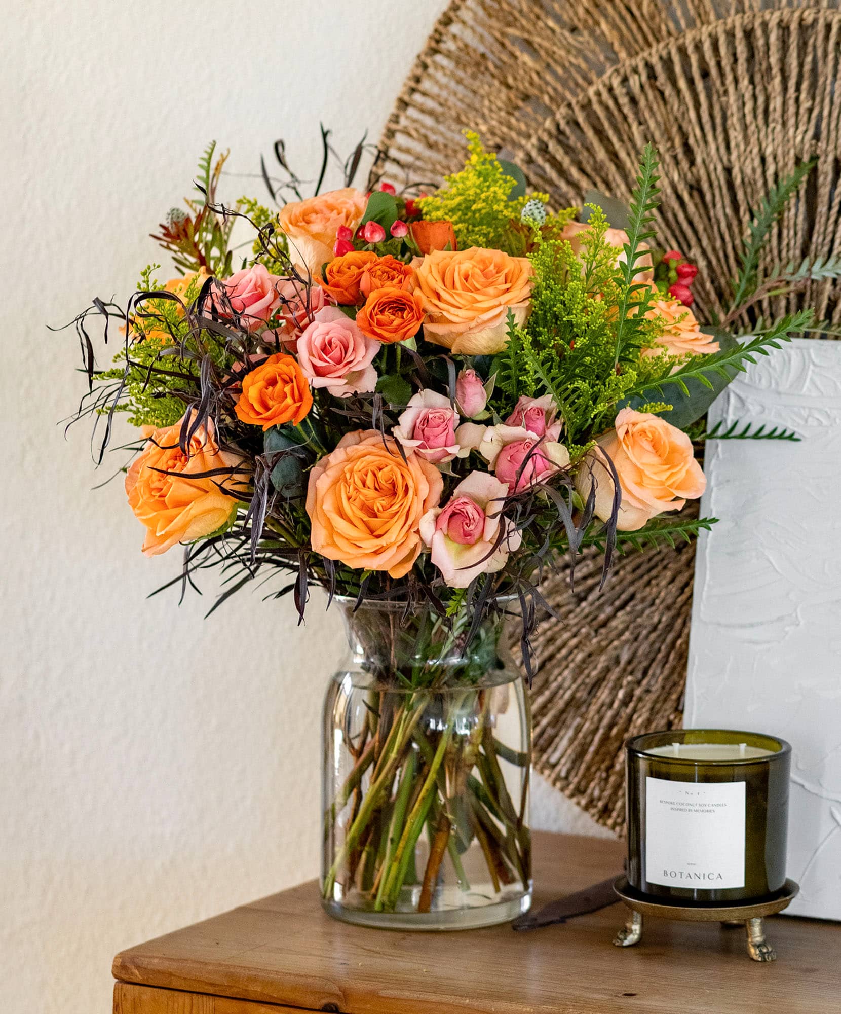 Vibrant floral arrangement of peach and pink roses with lush greenery in a glass vase.